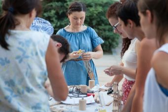 Plenerowy warsztat w ramach lekcji sztuki. Zdjęcie ukazuje kilku uczestników warsztatu, którzy z zaangażowaniem ścierają, tną drewno oraz sprawdzają efekty swojej pracy.