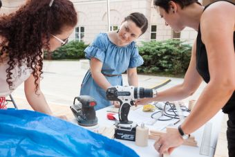Plenerowy warsztat w ramach lekcji sztuki. Plac Litewski, trzy kobiety, uczestniczące w wydarzeniu. Konsultują się, wykonując przy tym prace w drewnie.