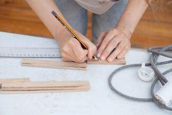 Plenerowy warsztat w ramach lekcji sztuki. Zbliżenie na damskie dłonie, nanoszące ołówkiem wymiary na drewnianą deskę.