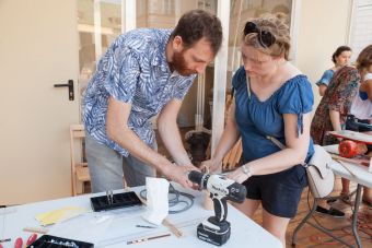 Zdjęcie ukazuje prowadzącego i uczestniczkę. Pochylają się oni nad drewnianymi elementami, prowadzący tłumaczy kobiecie kolejne etapy pracy. Na stole, przy którym stoją leży wiertarko-wkrętarka i drobne przyrządy do obróbki drewna.