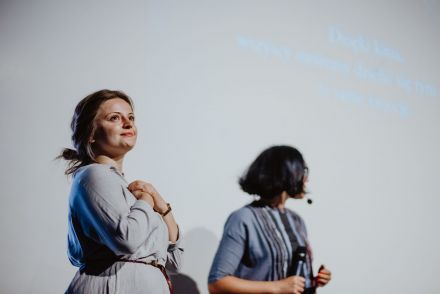 Kobieta się uśmiecha i układa ręce na sercu. W tle inna kobieta z mikrofonem.