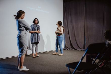 Trzy prowadzące stoją przed ekranem. Rzutnik powoli zaczyna się włączać.