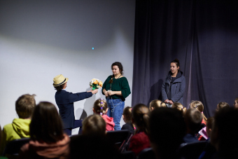 Klub filmowy dla najmłodszych w ramach Magicznej Latarni. Na zdjęciu ponownie ten sam duet aktorski, tym razem towarzyszy im mężczyzna, aktor Adam Organista. Na głowie ma kapelusz. Klęczy on przed prowadzącą i wręcza jej bukiet kwiatów. Obie kobiety mają zaskoczone miny.