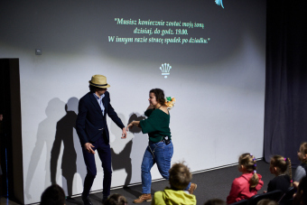 Klub filmowy dla najmłodszych w ramach Magicznej Latarni. Zdjęcie ukazuje scenę końcową przedstawienia wprowadzającego do seansu filmowego. Rozzłoszczona prowadząca usiłuje uderzyć mężczyznę bukietem kwiatów.  Na ścianie w tle ciągle widać napis "Musisz koniecznie zostać moją żoną dzisiaj, do godz. 19:00. W innym razie stracę spadek po dziadku".