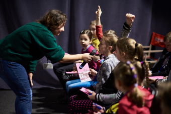 Klub filmowy dla najmłodszych w ramach Magicznej Latarni. Na zdjęciu  prowadząca ponownie przybliża mikrofon do innej dziewczynki z pierwszego rzędu.