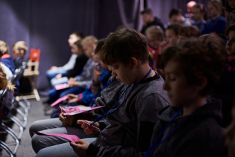 Klub filmowy dla najmłodszych w ramach Magicznej Latarni. Zbliżenie na jeden z rzędów krzesełek, na których siedzą dzieci. Na pierwszym planie widać chłopca przyglądającego się identyfikatorowi na swojej szyi.