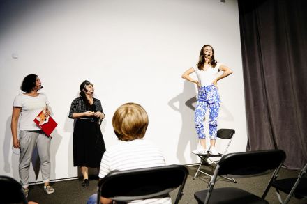 Kobieta stoi na krześle i mówi do dzieci przez mikrofon. Obok stoją dwie pozostałe animatorki.