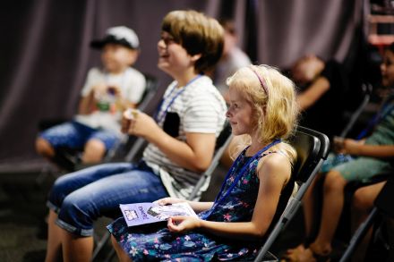 Dzieci siedzące na widowni. Dziewczynka trzyma na kolanach książeczkę zapowiadającą wydarzenie.