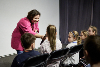 Zdjęcie ukazuje przybyłe dzieci oraz koordynatorkę, nawiązującą interakcję z publicznością. Trzyma mikrofon w ręce i przybliża go dziewczynce z widowni, oddając jej głos.