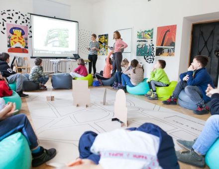 Na zdjęciu widać dzieci siedzące w okręgu i przyglądające się prezentacji. Dwie prowadzące opowiadają o architekturze. Na środku znajduje się wielkoformatowa tekturowa makieta miasta. Na zdjęciu widać dzieci siedzące w okręgu i przyglądające się prezentacji. Dwie prowadzące opowiadają o architekturze. Na środku znajduje się wielkoformatowa tekturowa makieta miasta.