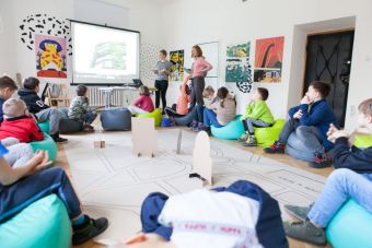 Na zdjęciu widać dzieci siedzące w okręgu i przyglądające się prezentacji. Dwie prowadzące opowiadają o architekturze. Na środku znajduje się wielkoformatowa tekturowa makieta miasta.