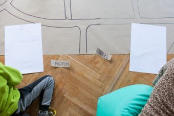 MalZdjęcie robione z góry. Widać  na nim fragment makiety, dwie białe kartki z naniesionymi szkicami oraz karteczki z napisem muzeum oraz hotel. Widać zarys dzieci siedzących obok.