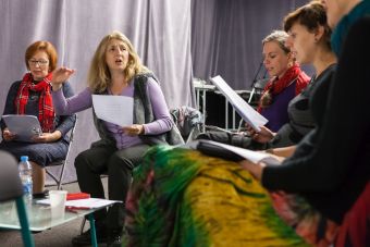 Na zdjęciu widzimy prowadzącą, która uczy śpiewu oraz uczestniczki. Wszystkie zgromadzone kobiety siedzą, trzymają kartki w dłoniach i śpiewają.