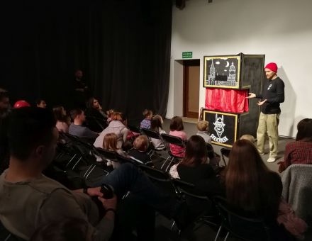 Zdjęcie przedstawia publiczność i scenę ze stojącym na niej mężczyzną w czerwonej czapce wskazującego rękami na scenę teatru pacynkowego. Zdjęcie przedstawia publiczność i scenę ze stojącym na niej mężczyzną w czerwonej czapce wskazującego rękami na scenę teatru pacynkowego.