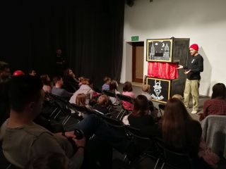 Zdjęcie przedstawia publiczność i scenę ze stojącym na niej mężczyzną w czerwonej czapce wskazującego rękami na scenę teatru pacynkowego.