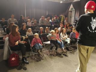 Grupa dzieci wpatrzona jest w scenę, na której stoi aktor.