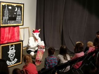 Koziołek Skejciołek kuca przed publicznością opierając się o deskorolkę.