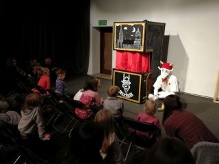 Koziołek Skejciołek wraz ze swoimi scenicznymi atrybutami i  sceną z kurtyną siedzi przed gronem publiczności. Są to dzieci i ich opiekunowie.