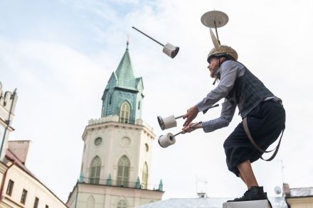 Mężczyzna stoi na dachu kamienicy. Na głowie ma czapkę wikinga z zamontowanym do niej talerzem. Żongluje trzema pałeczkami.