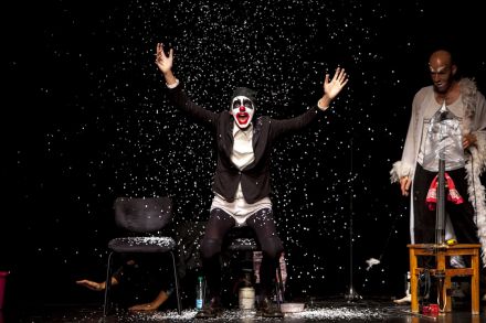 Mężczyzna ubrany na czarno, z twarzą pomalowaną jak clown, wstaje z krzesła. Otacza go biały pył. Z jego prawej strony stoi drugi mężczyzna.