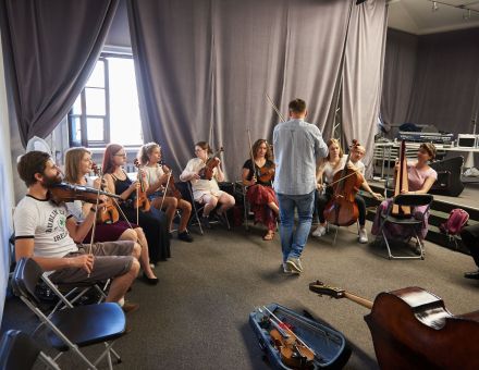 Na zdjęciu widzimy salę pełną uczestników, mających przy sobie instrumenty muzyczne, na środku stoi mężczyzna ze skrzypcami. Na zdjęciu widzimy salę pełną uczestników, mających przy sobie instrumenty muzyczne, na środku stoi mężczyzna ze skrzypcami.