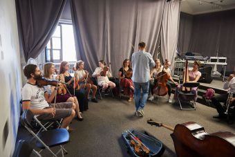 Na zdjęciu widzimy salę pełną uczestników, mających przy sobie instrumenty muzyczne, na środku stoi mężczyzna ze skrzypcami.