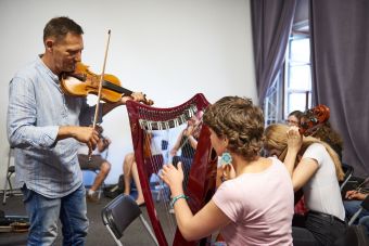 Na pierwszym planie widzimy mężczyznę grającego na skrzypcach oraz kobietę grającą na harfie. W tle pozostali uczestnicy.
