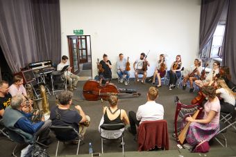 Na zdjęciu widać wszystkich uczestników siedzących w okręgu. Mają przy sobie instrumenty, na środku leży kontrabas.