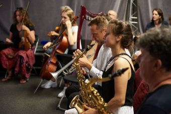 Zbliżenie na kilku uczestników podczas grania. Na pierwszym planie widzimy  mężczyznę i kobietę z saksofonem oraz mężczyznę grającego na trąbce. W oddali kobieta grająca na wiolonczeli oraz dwie skrzypaczki.