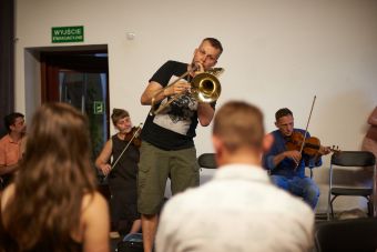 Na zdjęciu widzimy grającego na trąbce prowadzącego. Stoi on na środku. Reszta siedzi nadal w okręgu i również gra na instrumentach.