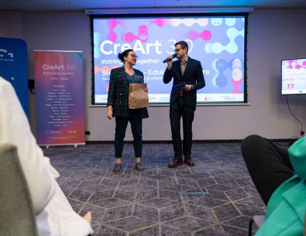 Kobieta i mężczyzna przemawiają do publiczności. Śmieją się przy tym.