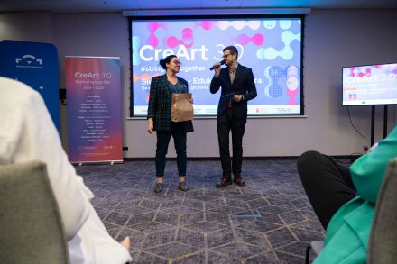 Kobieta i mężczyzna przemawiają do publiczności. Śmieją się przy tym.