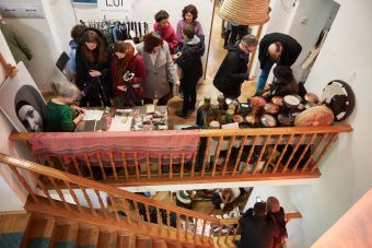 Na zdjęciu widać zgromadzonych ludzi, wystawców oraz dwa stoiska. Na jednym znajdują się ręcznie wykonane instrumenty, a na drugim sprzedawane są akcesoria typu woreczki na zakupy, notesy wykonane przez uchodźczynie ]