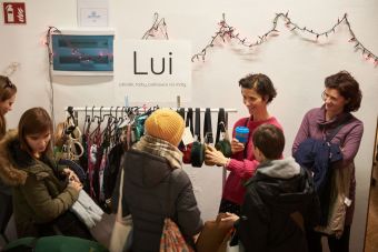 Na zdjęciu widać stoisko z plecakami, torbami, pokrowcami na maty firmy Lui. Sprzedająca rozmawia z zainteresowanymi ofertą osobami.