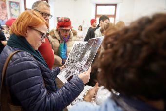 Zdjęcie ukazuje stoisko z grafikami ( kartki, plakaty) których głównym tematem jest Lublin. Jedna z przybyłych osób trzyma w rękach dwa plakaty. Pierwszy przedstawia Bramę Krakowską, a drugi Stare Miasto w Lublinie.