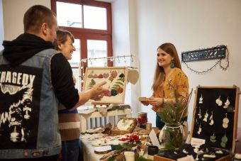 Zdjęcie ukazuje sprzedającą przy stoisku z biżuterią i ozdobami. Prowadzi ona rozmowę z klientami.