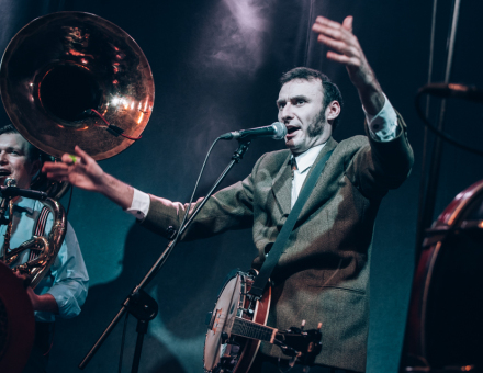 Na pierwszym planie muzyk z  banjo, rozkładając dłonie śpiewa do mikrofonu. W tle muzyk z tubą.