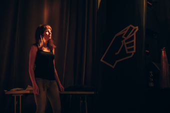 Młoda dziewczyna stoi na tle ciemnej tkaniny z logotypem zespołu Hańba czyli zaciśniętą pięścią, wpatruje się w scenę.  Światła sceny rozjaśniają jej twarz.