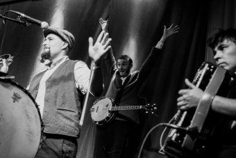 Czarno białe zdjęcie przedstawiające trzech muzyków zespołu Hańba. Dwóch z nich unosi ręce do góry, trzeci trzyma akordeon na którym gra.