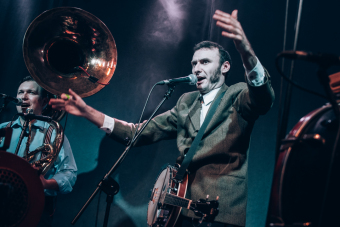 Na pierwszym planie muzyk z  banjo, rozkładając dłonie śpiewa do mikrofonu. W tle muzyk z tubą.