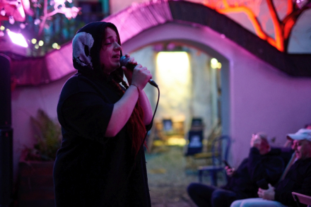 Wokalistka ma zamknięte oczy i kaptur na głowie. Śpiewa do mikrofonu.