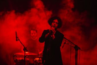 Wokalista zespołu Alpha Strategy na środku fotografii stoi przy mikrofonie z przymkniętymi oczami, w tle dym i ostre czerwone światło reflektora. Na drugim planie perkusista.