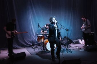 Widok na cały zespół Alpha Strategy stojący na scenie na tle ciemnej tkaniny.  Widzimy instrumenty perkusyjne, gitarę elektryczną, basową i wokalistę.