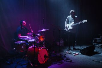 Muzycy zespołu Alpha Strategy podczas koncertu. Na zdjęciu perkusista i basista. Perkusista oświetlony jest fioletowo-różowym światłe, basista  światłem niebiesko-sinym.