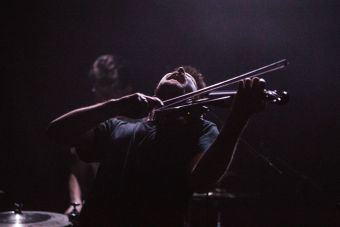 Portret skrzypka zespołu Ciśnienie stojącego na scenie. Mężczyzna gra na instrumencie, ma odchyloną do tyłu głowę. Za nim niewyraźna sylwetka perkusisty. Ciemne tło.