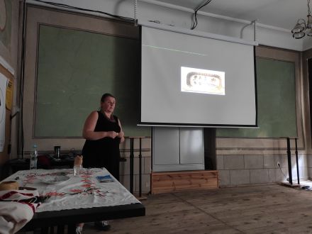 Na ekranie jest wyświetlona prezentacja, obok stoi prowadząca oraz stół z przedmiotami ludowymi. 