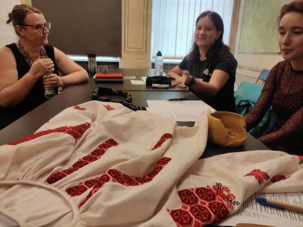 Na stole leży wyhaftowana koszula, na którą przyglądają się kobiety.