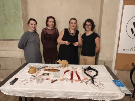 Cztery kobiety stoją przed stołem, na którym znajdują się elementy ubioru ludowego, rzeźby oraz naczynia. Przedmioty te stoją na haftowanym obrusie. 