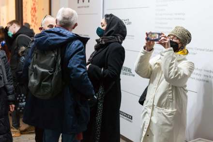 Ludzie stoją w grupkach i rozmawiają. Jedna kobieta robi zdjęcie.