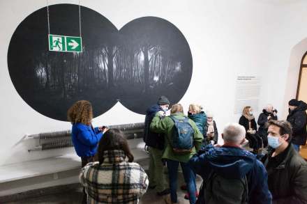 Na białej ścianie narysowany jest widok z lornetki nocą. Przedstawia on las. Ludzie stoją przed ścianą i oglądają to dzieło.